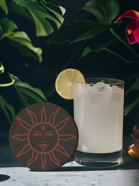 Round ceramic coaster in deep black featuring an intricate burnt-orange sun illustration with a serene, symmetrical face, radiating curved rays, and a delicate star detail on the forehead. The smooth, matte surface enhances the contrast between the dark background and the warm-toned design, creating a celestial, vintage-inspired aesthetic.